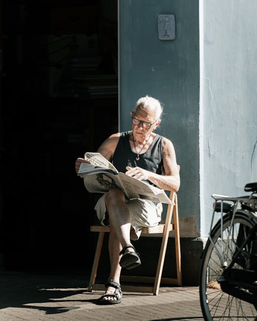 An elderly woman with short gray hair and glasses is seated outdoors on a wooden chair in front of a light blue wall with a small electrical box. She is wearing a black sleeveless top, beige capris, a
